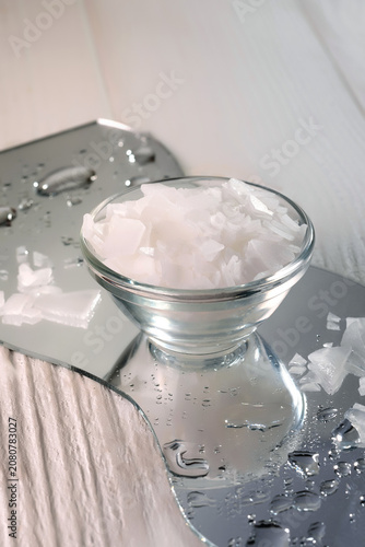 Magnesium bath salt flakes in a small glass bowl on a wet mirror surface with water drops, spa and wellness concept