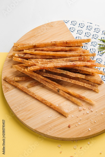 Dried fish sticks snack served on a round wooden cutting board with sea salt crystals, bright background