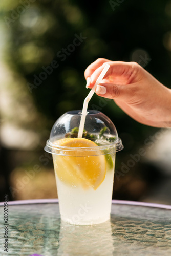 Refreshing summer lemonade with lemon and lime slices in a plastic takeaway cup with a straw, outdoors background