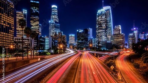 Night city lights blaze above busy highway. Cars streak red and white through urban arteries. Skyscrapers glow with energy, windows shine bright. Traffic flows endlessly under starless, dark sky