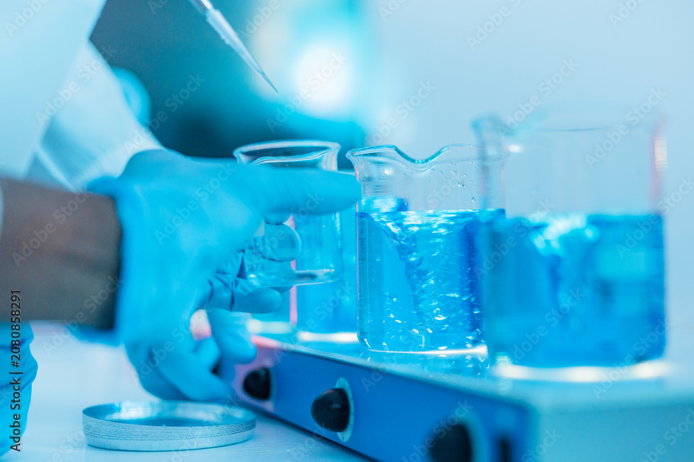 Fototapeta premium Gloved Lab Technician Positions Glass Beakers Filled with Vivid Blue Solution on Stirring Platform 