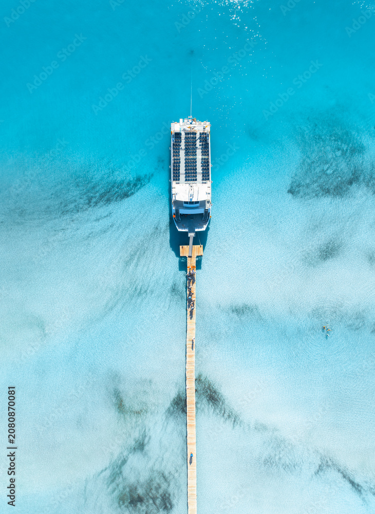 Fototapeta premium Top down aerial view of passenger ship yacht at wooden pier in Mallorca, Spain. Cruise boat docked at jetty in clear turquoise sea water near sandy beach. Drone tropical exotic summer landscape