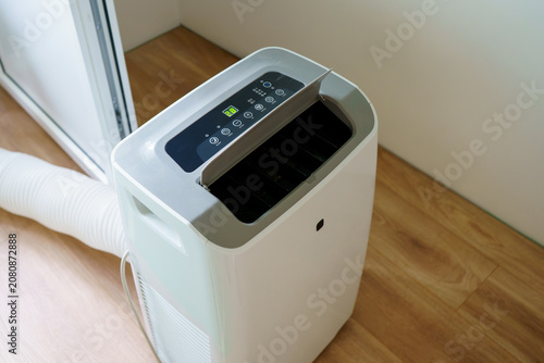 A modern portable mobile air conditioner unit stands on a wooden floor during a hot summer day, controlling temperature and relative humidity inside the room.