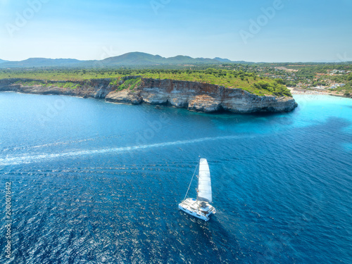 Aerial view of sailing catamaran yacht in turquoise sea water bay. Sailboat. Mallorca, Spain. Luxury boat cruising in ocean cove near sandy beach, resort houses on rocky coast. Drone summer landscape