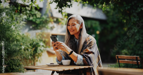 Schilderij op canvas A silver haired mature woman, wrapped in a plaid shawl sits alone at a wooden outdoor cafe table and smiles warmly at her smartphone screen in bright golden sunlight