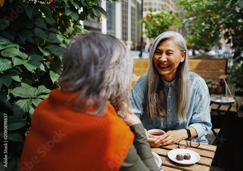 Konstfotografi A long silver haired woman in a denim shirt, bursts into genuine laughter, while holding a cup of tea across a wooden table from a companion in an orange wrap