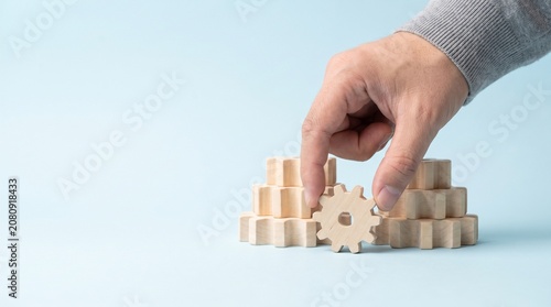 Hand placing a wooden gear into a stack of gears, representing business process, teamwork, and integration concepts on a blue background.
