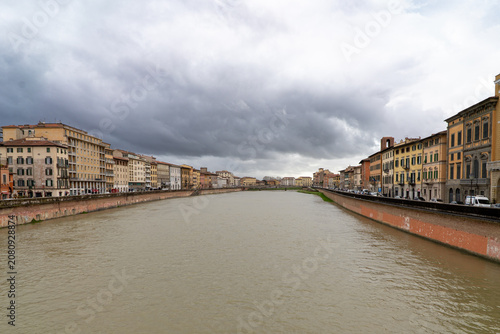 View of the Arno river flowing through Pisa, Italy, with colorful historic buildings lining the banks under a dramatic cloudy sky