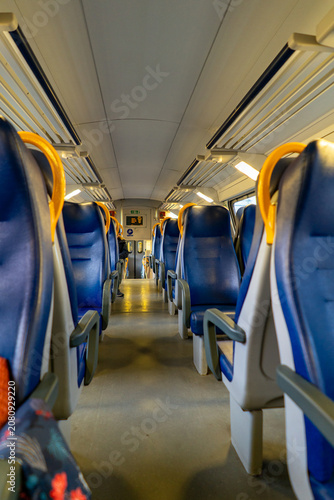 The perspective shot shows rows of empty blue seats with yellow grab handles inside a passenger train carriage, illuminated by overhead lights