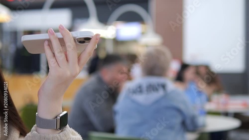 Hand holds phone, capturing blurred cafe scene with people. Person wears watch, focused on screen. Background shows casual dining, soft lighting. Ideal for social media, travel. Suitable for lifestyle
