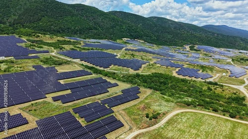 Rows of solar panels cover the ground in a vast area near hills. The sky is blue with some clouds. This site shows the use of solar energy for power generation.