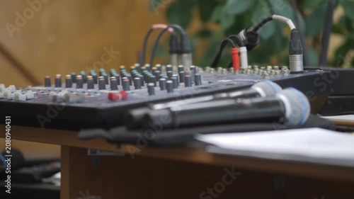 Audio mixer and microphones sit on wooden table. Cables connect devices to mixing board. Background shows green plants and warm lighting. Ideal for music production, live events, or studio setups