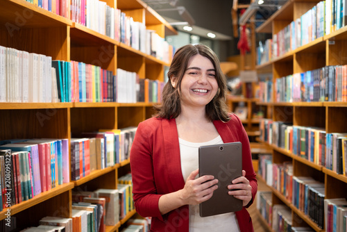 Smiling White Latin Employee in Red Blazer Holding Tablet Standing in Bookstore Library Aisle Wide Medium Shot
