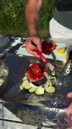 Close up of hands placing red bell pepper slices on top of seasoned fish at campsite.