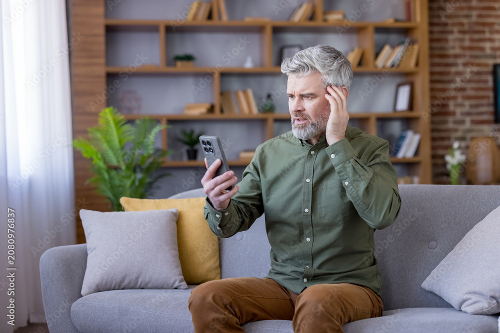 Fototapeta premium Mature man with a beard sitting on a sofa in his living room, looking at his smartphone with a distressed expression after reading upsetting information or news