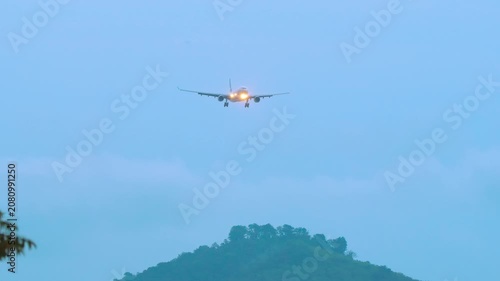 Passenger jet airplane descending for landing approach with bright headlights on over a green tropical hill during blue hour twilight, telephoto front shot.