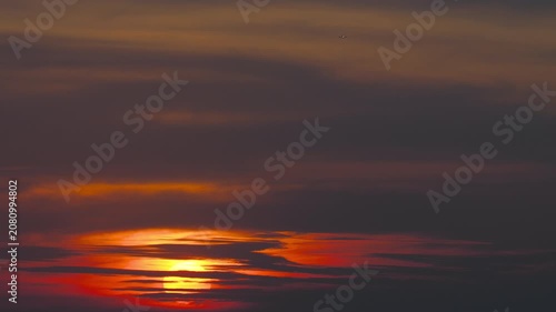 Sunset shining through dark clouds with a tiny airplane flying in the distance under a dramatic orange and red evening sky, real time telephoto shot