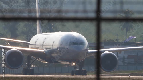 Passenger airplane taxiing on runway at tropical airport seen through blurred fence grid with palm trees in sunny weather, telephoto close-up shot with heat haze distortion