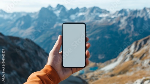 Subject A hand holding a smartphone with a blankwhite screen. Background Majestic, snow-capped, jagged mountains under a clearbright sky. The background is slightly out of focus (bokeh