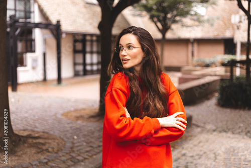 Thoughtful woman with long brunette hair wearing orange sweatshirt and glasses, looking away with curious expression, arms crossed in park, outdoor portrait, modern lifestyle, casual fashion, thinking