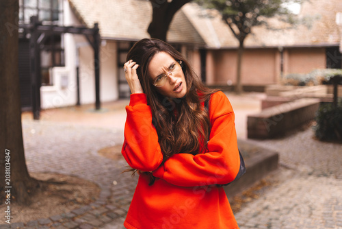 Pensive young woman in red sweater holding hand on head looking thoughtfully upwards and to the side in contemplation of a problem or decision while outdoors on urban street background