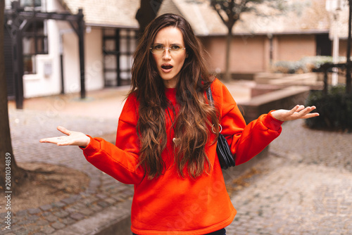 Confused woman in red sweater looking at camera with shocked facial expression and shrugging shoulders with open palms gesture outdoors expressing uncertainty disbelief cluelessness misunderstanding