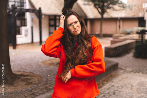 Confused young woman in red sweater holding hand on head looking puzzled and uncertain with doubtful expression outdoors on urban street background