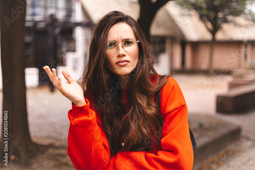 Confused young woman in red sweater questioning something with a puzzled and bewildered facial expression outdoors raising hand in a gesture of uncertainty and misunderstanding