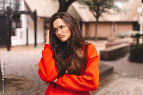 Pensive young woman in red sweater holding hand on head looking thoughtfully upwards and to the side in contemplation of a problem or decision while outdoors on urban street background