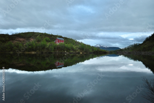 Mountain lake in Norway with mirror reflections of surrounding peaks, forests, clouds, and a small red cabin on the shore during a calm, overcast evening.