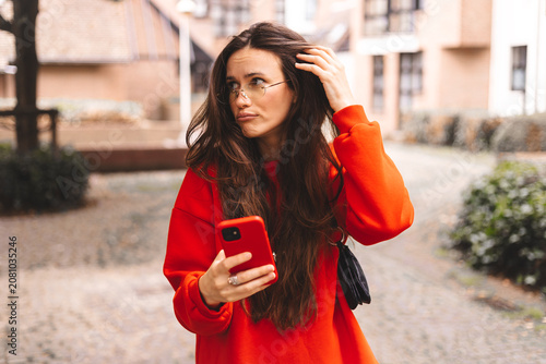 Quadro su tela Confused young woman in eyeglasses holding smartphone and touching long hair wit