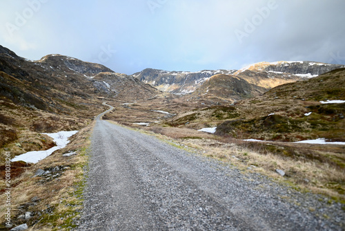 Photography Gravel road stretching into the distance through the mountains of Norway in late spring, surrounded by dry grass and small patches of lingering snow