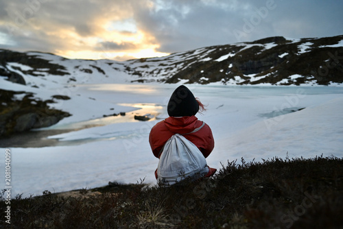 Woman in an orange rain jacket sitting with her back to the camera by a snowy mountain lake in Norway at sunset, surrounded by dramatic alpine peaks.