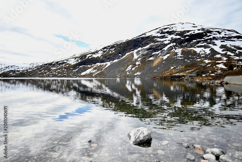 Mountain lake in Norway with gentle ripples reflecting peaks lightly covered in snow during late spring, surrounded by serene Nordic scenery.