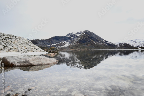 Mountain lake in Norway reflecting clouds and snow-dusted peaks in late spring, with gentle ripples adding texture to the tranquil water surface.