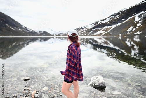 Woman in a pink-purple shirt and cap standing with her back to the camera by a mountain lake in Norway in May, with lightly snow-covered peaks behind.