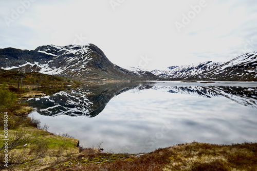 Mountain lake above a Norwegian fjord with mirror reflections of peaks lightly covered in snow during late spring.