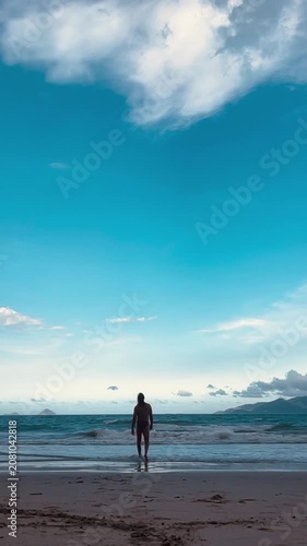 Solitary silhouette standing at shoreline beneath vast teal sky with billowy white cloud and gentle surf, with ocean, shoreline, silhouette, person detail, showing standing, beach, horizon, sky