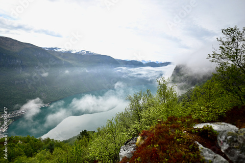 Aerial view of a Norwegian fjord with turquoise water, dramatic rocky cliffs, and low clouds drifting over the surface.