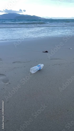 Discarded plastic bottle drifting across wet sand beside incoming tide on quiet empty shoreline under overcast sky, with beach, sand, bottle, litter detail, showing shore, ocean, wave, sea context