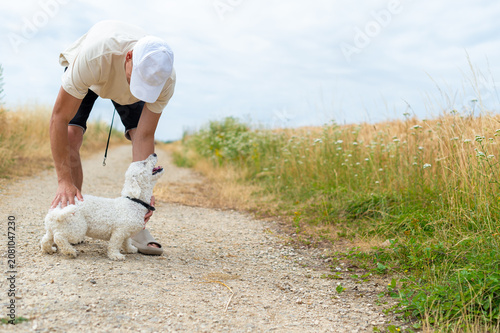 Foto A man wearing a white cap bends over to pet his small white Bichon Frise dog on a gravel country road