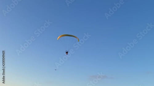 A powered paraglider pilot with a yellow canopy flies over the beach against an orange sunset.
