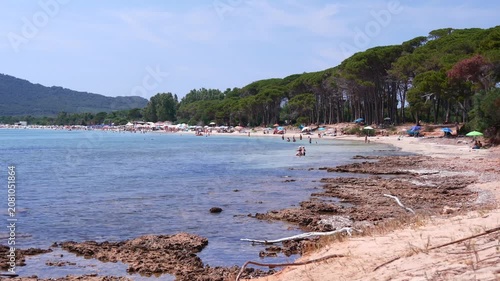 Sunny coastal view of Spiaggia di Mugoni in Sardinia, with clear blue water, rocky shoreline, and pine-covered hills beyond.