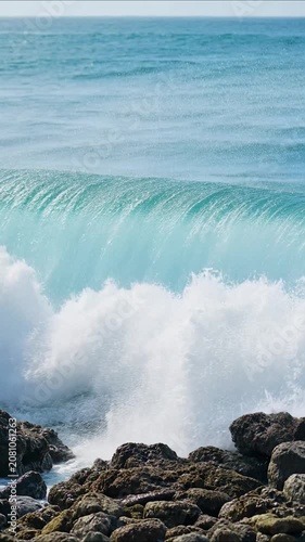 Powerful turquoise ocean wave crashing on a rocky shore during a sunny day with clear blue sky in the background