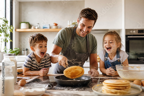 Pai e filhos pequenos fazendo panquecas juntos em uma cozinha ensolarada - Conceito de diversão em família, café da manhã especial e amor paterno.