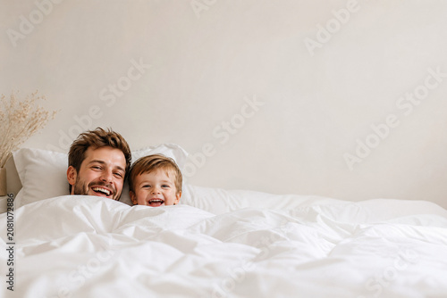 Pai e filho pequeno rindo sob lençóis brancos em cama confortável com amplo espaço para texto - Conceito de intimidade familiar, manhã feliz e amor incondicional.