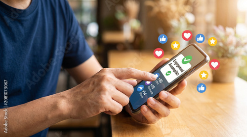A person holds a mobile device displaying social media engagement metrics featuring colorful floating reaction icons like hearts and thumbs up in a bright indoor setting