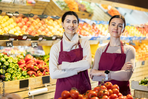 Фотография Two female seller of fruit and vegetable store waits for visitors near counter and greets clients