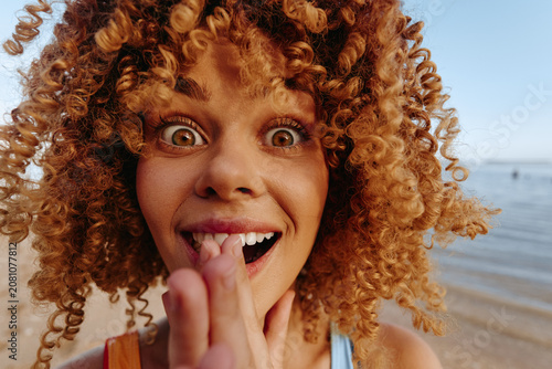 Fotografija Close-up of a young woman with curly hair on a sunny beach, playfully sticking o