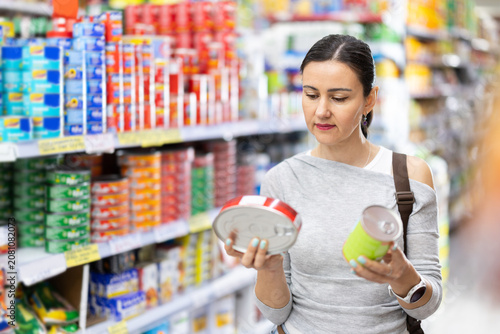 Woman buyer examines display case, reads product label information before buy canned food. Client compares different options item, chooses product goods and check shelf life